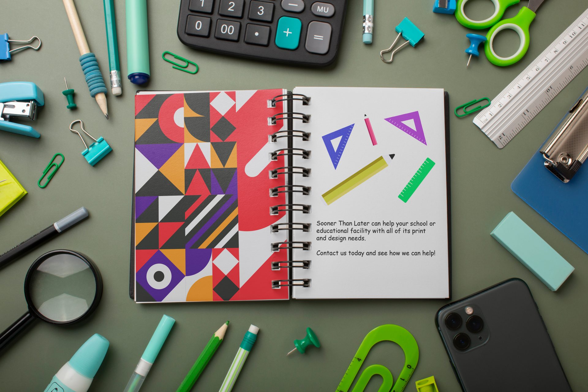 Spiral-bound notebook with a colourful geometric printed cover, styled on a desk with stationery including pens, scissors, paper clips and a calculator